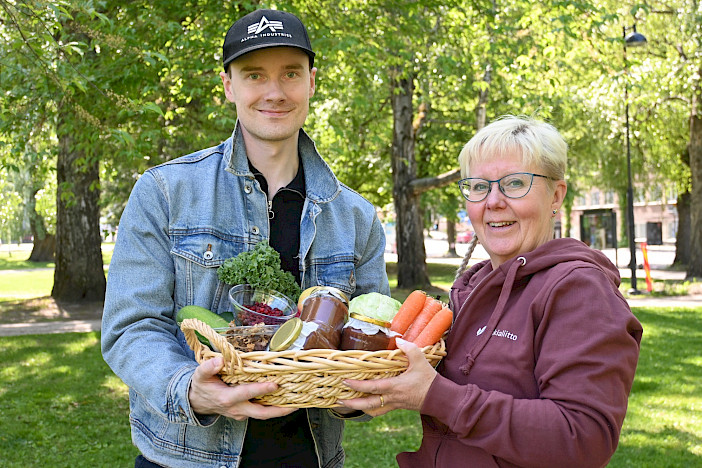 Keliakialiiton ravitsemusasiantuntija Miika Wynne-Ellis ja HoReCa-asiantuntija Eija Lindberg pitelevät koria, jossa on juureksia, sieniä, puolukoita ja omenasosepurkkeja. Kuva: Tuulamaria Lempiälä.